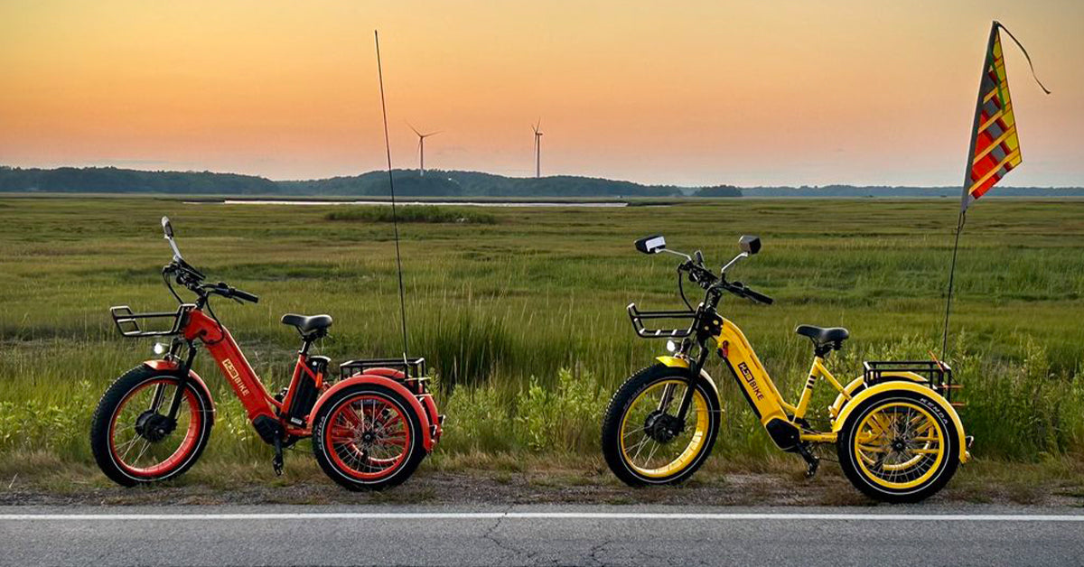 Red and yellow HJM TriHauler electric trikes beside coastal grassland at sunset